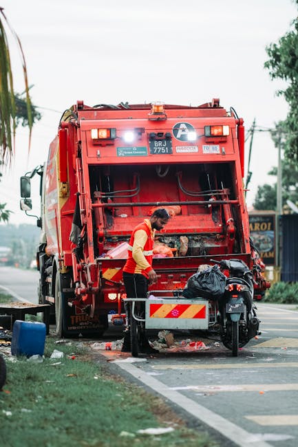 A worker operating a large red waste collection vehicle on a roadside during the daytime, with the rear compartment open for waste collection. The worker, wearing a high-visibility yellow and red vest