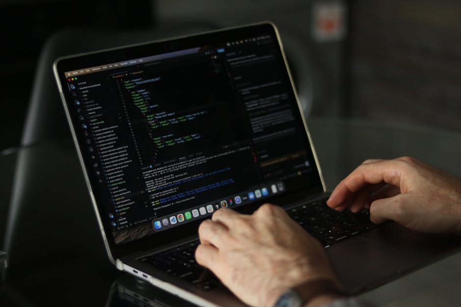 A close-up view of a person's right hand using a laptop computer placed on a wooden surface. The hand is positioned over the keyboard, with fingers hovering above the keys. The individual is wearing a