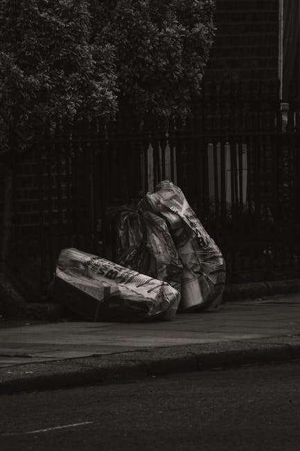 A large, dirty beige or light brown fabric sack filled with miscellaneous waste materials rests on the uneven, asphalt pavement of a narrow urban street. The sack appears worn and stained, with loose 